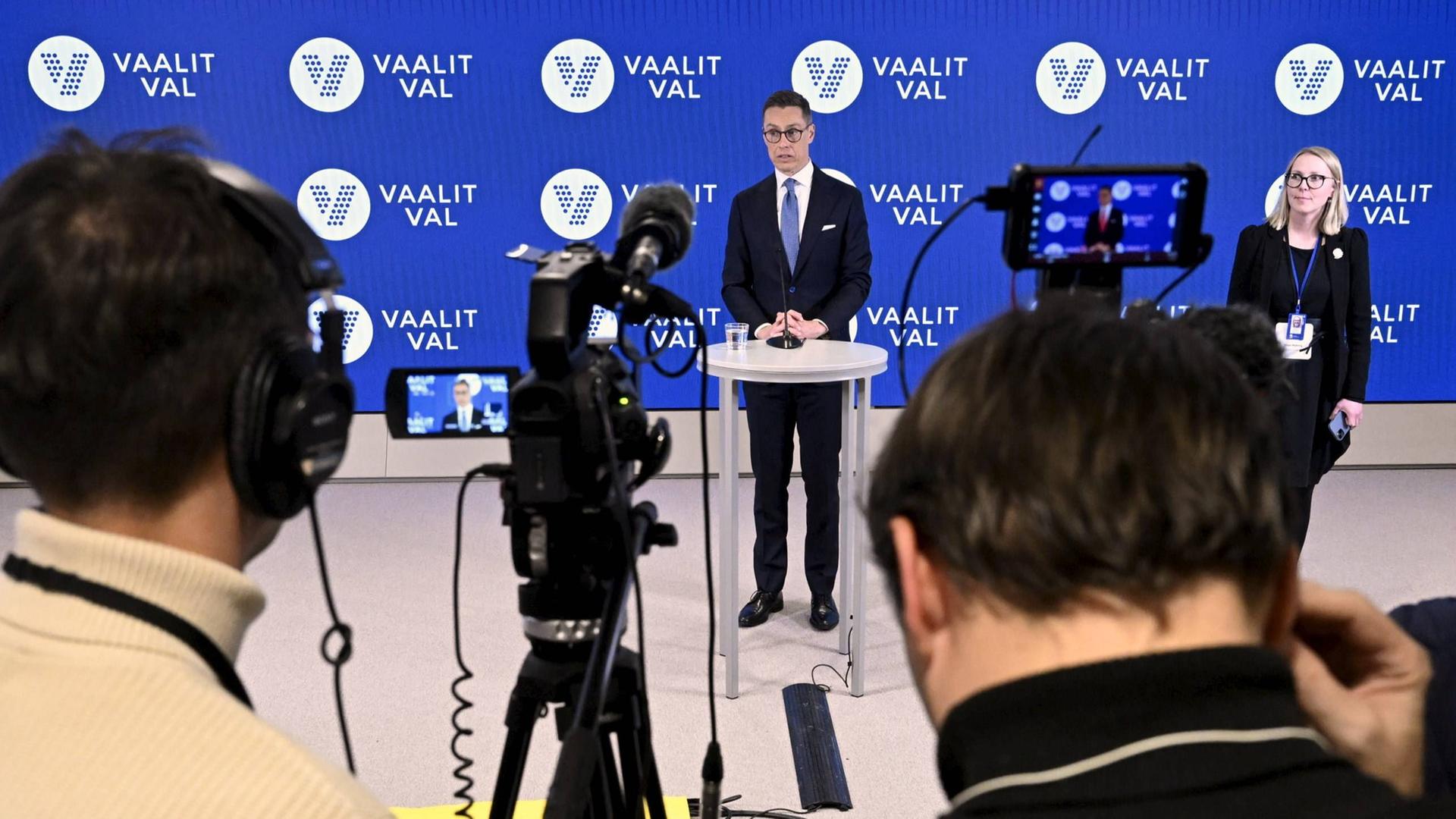 Wahlsieger Alexander Stubb steht auf einer Bühne vor einem Mikrofon; im Vordergrund sind zwei Jounalisten mit Kameras zu sehen.