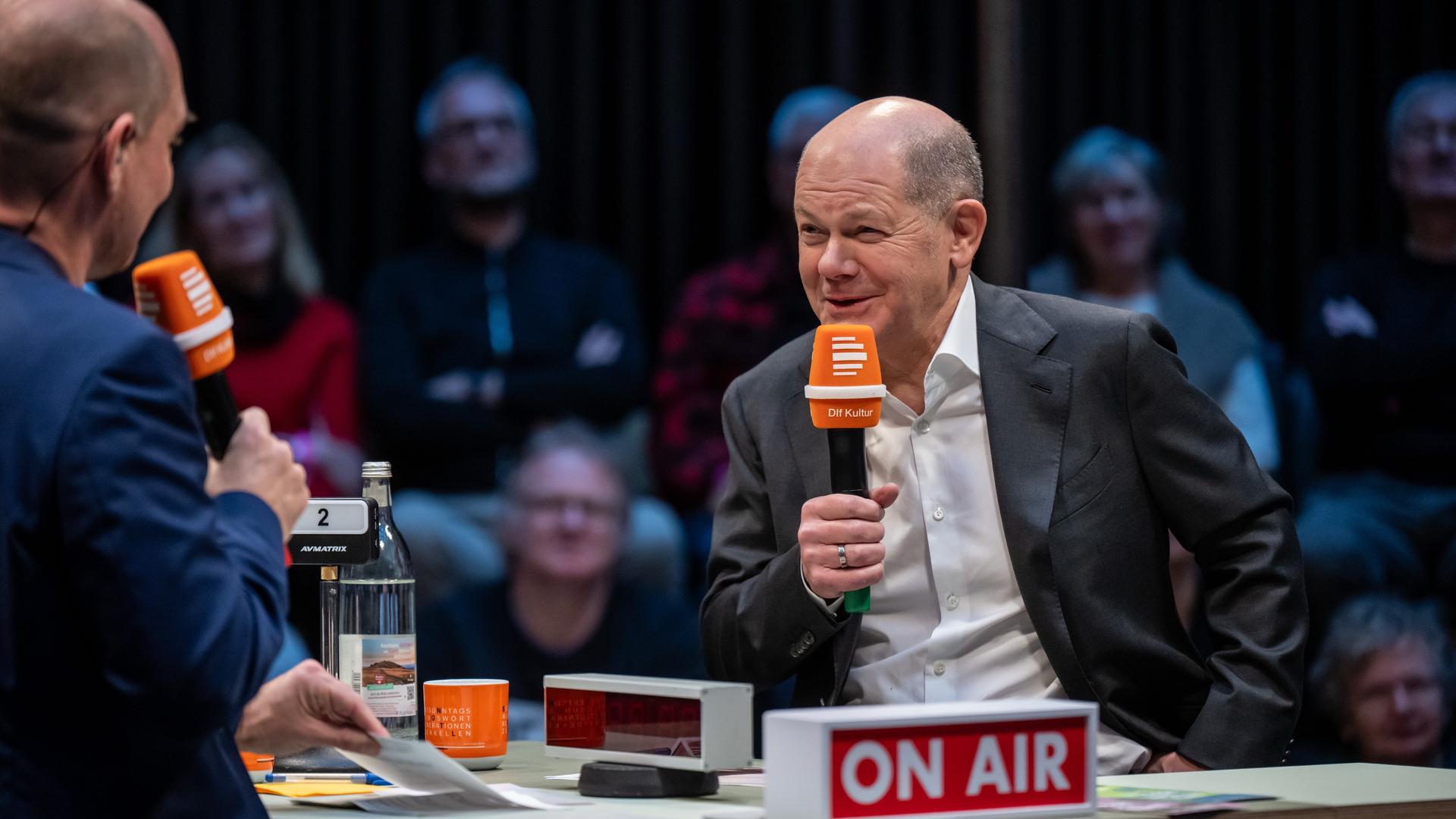 Bundeskanzler Olaf Scholz (SPD) sitzt am Tisch der Live-Radioshow "Studio 9 -. Der Tag mit" und blickt lachend zu Moderator Korbinian Frenzel.