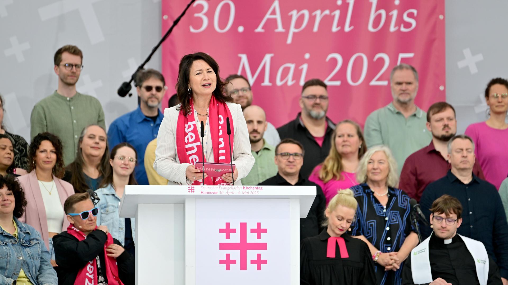 Anja Siegesmund steht mit dem Kirchentags-Schal um den Hals an einem Rednerpult, im Hintergrund viele Menschen, teilweise in Priestergewändern. Anja Siegesmund steht mit dem Kirchentags-Schal um den Hals an einem Rednerpult, im Hintergrund viele Menschen, teilweise in Priestergewändern.