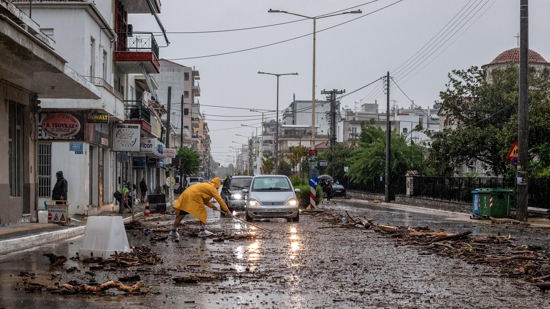 Autos fahren auf einer halb überschwemmten Straße, von deren Seiten ein Mann in gelbem Regenmantel versucht Äste und angeschwemmte Gegenstände zu bergen.