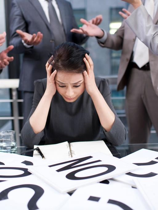 Eine Frau sitzt vor einem Tisch mit vielen Zetteln, auf denen Fragezeichen und das Wort "No" zu sehen ist. Sie stützt aus Überforderung ihren Kopf in die Hände.