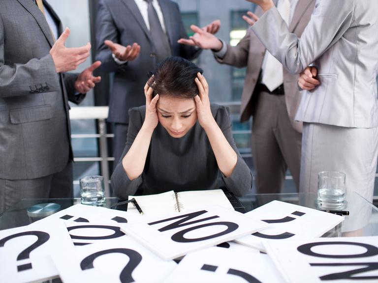 Eine Frau sitzt vor einem Tisch mit vielen Zetteln, auf denen Fragezeichen und das Wort "No" zu sehen ist. Sie stützt aus Überforderung ihren Kopf in die Hände.