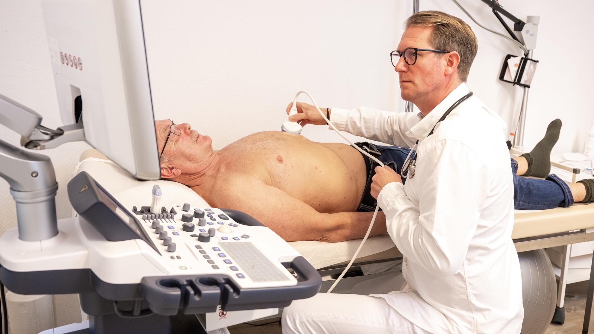 Ein Hausarzt hält ein Ultraschallgerät auf den Bauch eines älteren, oberkörperfreien Patienten. Dabei schaut der Arzt in den Monitor vor sich.