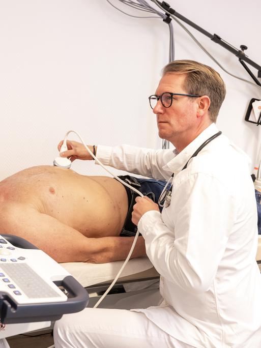 Ein Hausarzt hält ein Ultraschallgerät auf den Bauch eines älteren, oberkörperfreien Patienten. Dabei schaut der Arzt in den Monitor vor sich.