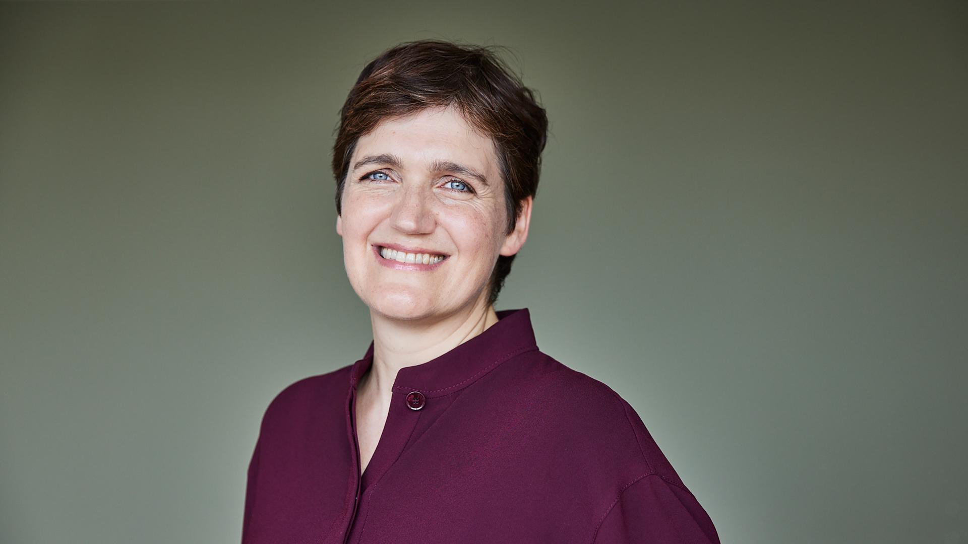 Katja Urbatsch trägt eine weinrote Bluse, steht vor einer graugrünen Wand und lächelt in die Kamera.