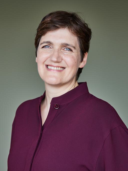 Katja Urbatsch trägt eine weinrote Bluse, steht vor einer graugrünen Wand und lächelt in die Kamera.