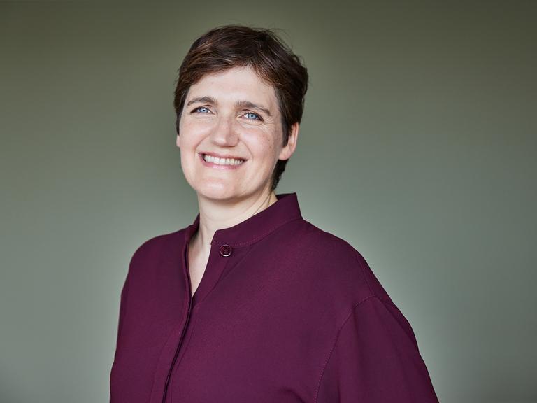 Katja Urbatsch trägt eine weinrote Bluse, steht vor einer graugrünen Wand und lächelt in die Kamera.