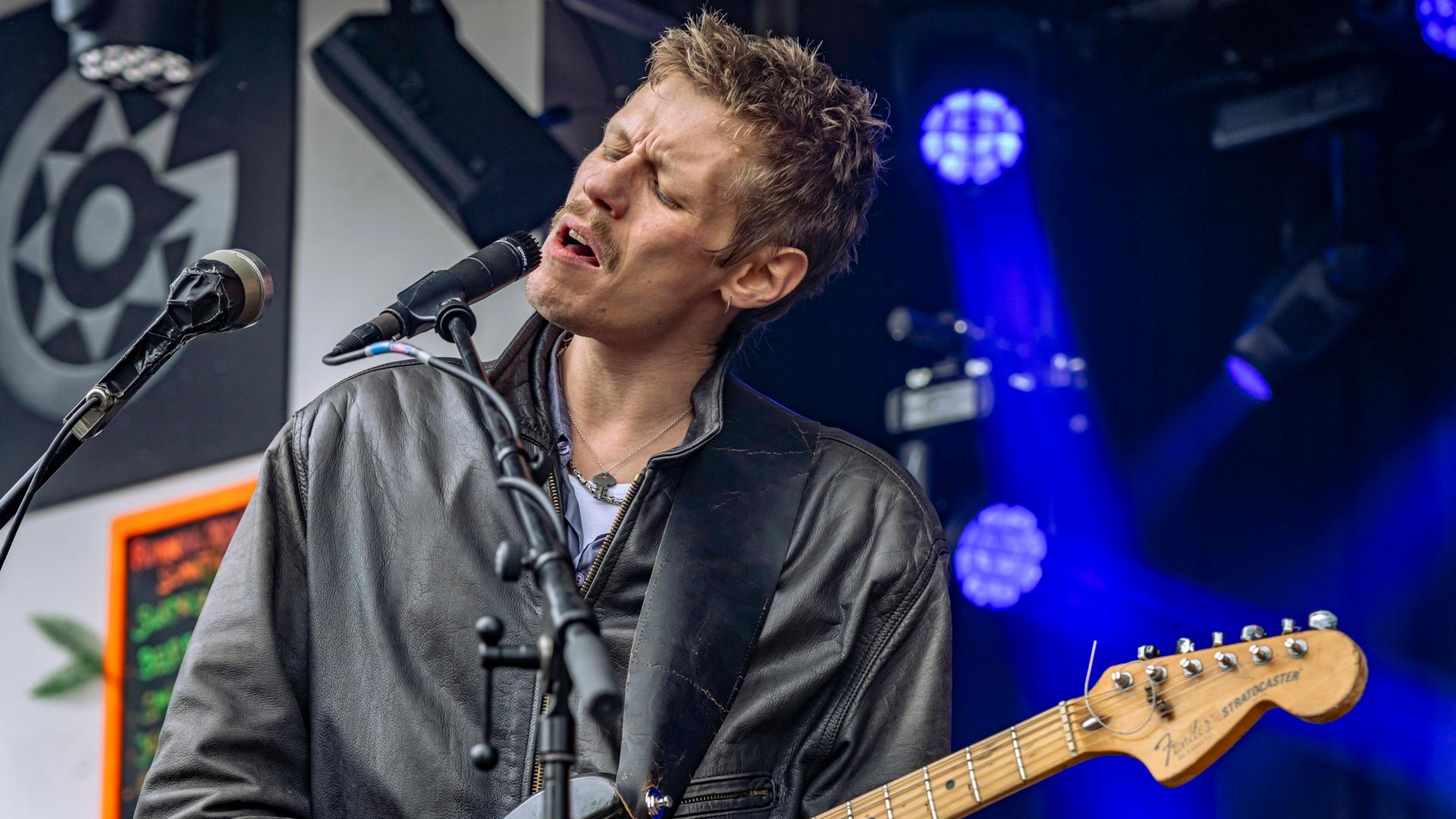 Wir sehen den Musiker Jesper Munk wie er gerade auf der Bühne steht, singt und Gitarre spielt.  Wir sehen den Musiker Jesper Munk wie er gerade auf der Bühne steht, singt und Gitarre spielt.