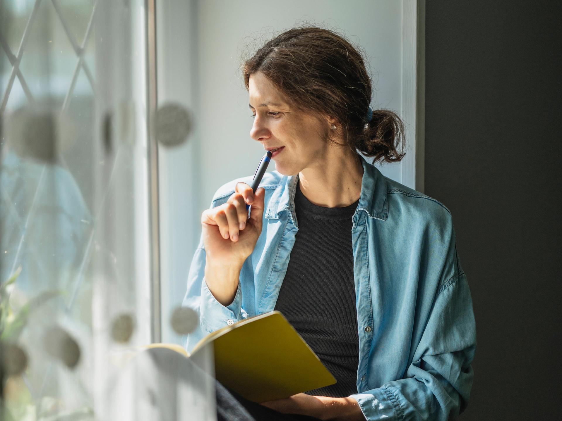 Eine Frau sitzt am Fenster mit einem Schreibheft und einem Stift und schaut gedankenvoll nach draussen.