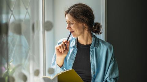 Eine Frau sitzt am Fenster mit einem Schreibheft und einem Stift und schaut gedankenvoll nach draussen.