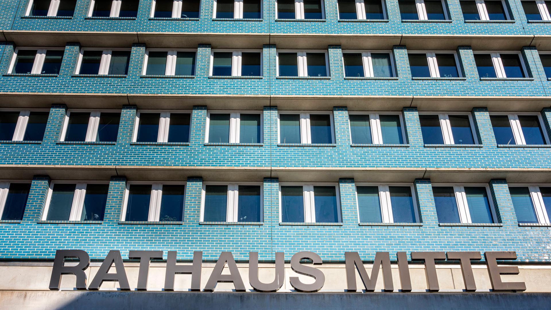 Ein mehrstöckiges Gebäude mit türkisfarbigen Wänden und vielen Fenstern, die gleichförmig angeordnet sind. Im Vordergrund   ist der Schriftzug "Rahtahus Mitte" zu sehen, der auf dem Vordach des Gebäudes steht. 