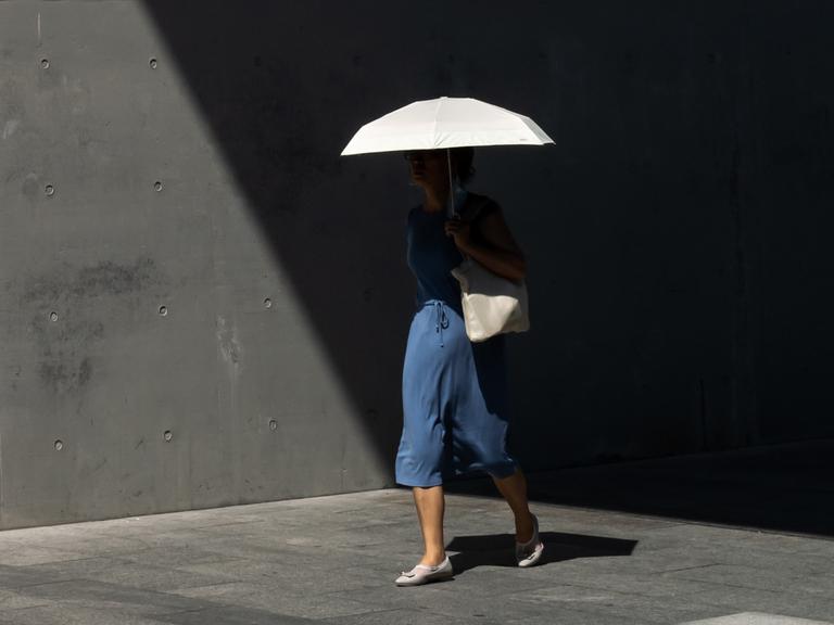Frau im blauen Kleid, mit weißer Umhängetasche und weißen Schuhen trägt eine weißen Schirm. Sie läuft aus dem Schatten heraus in die Sonne. Gehweg und Tunnelwand sind trist und grau. Ihr Oberkörper ist durch Schatten kaum zu erkennen.