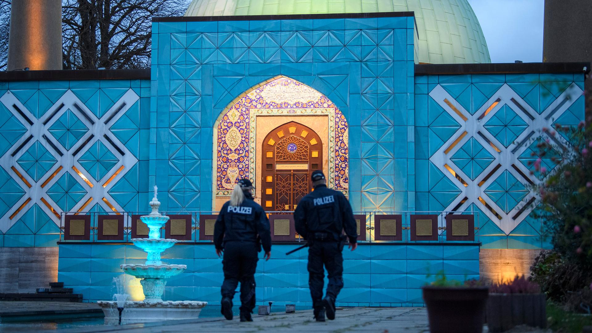 Zwei Polizisten stehen im Abendlicht vor der beleuchteten Imam-Ali-Moschee des Islamischen Zentrums Hamburg. 