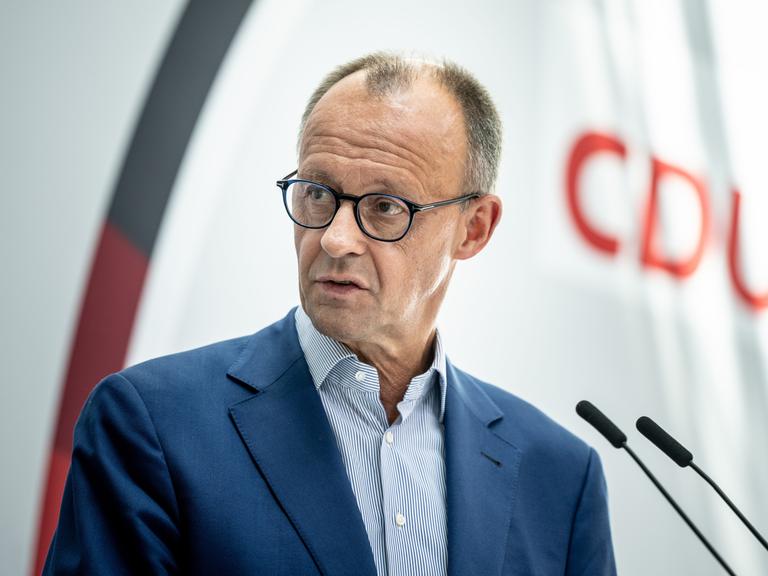 Ein Mann in den Sechzigern mit Brille, Friedrich Merz, spricht an einem Stehpult, im Vordergrund sind zwei Mikros zu erkennen, im Hintergrund der Schriftzug CDU.