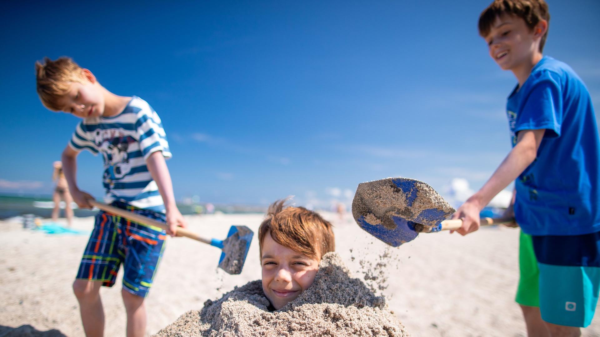 Das Foto zeigt drei spielende Jungen am Strand. Einer wird von den beiden anderen im Sand eingebuddelt. Das Foto zeigt drei spielende Jungen am Strand. Einer wird von den beiden anderen im Sand eingebuddelt.