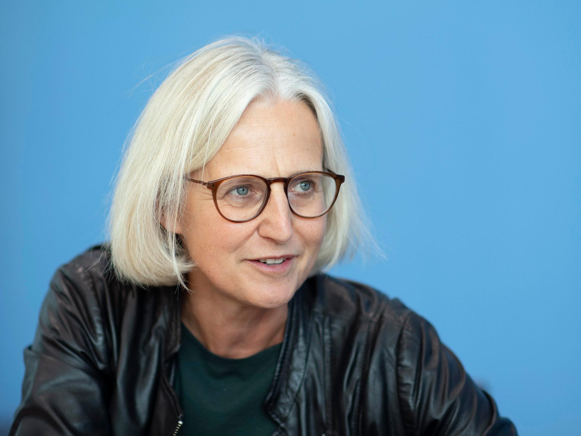 Die Autorin Christiane Hoffmann im Portrait von der Seite. Sie trägt eine Brille, hat blonde kurze Haare und trägt dunkle Kleidung. 