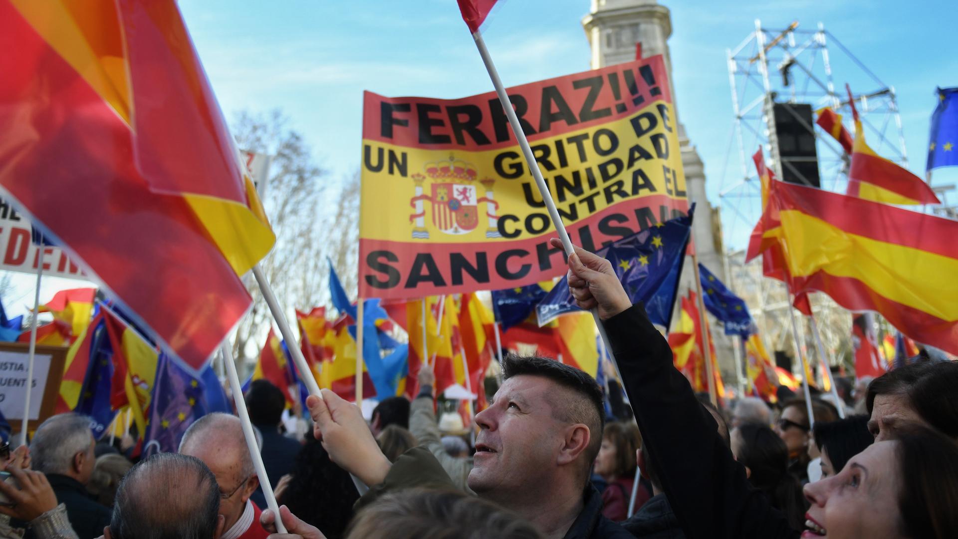 Viele Menschen schwenken rot-gelbe Flaggen und Transparente mit Aufschriften auf Spanisch. Der Himmel ist blau, die Sonne scheint.