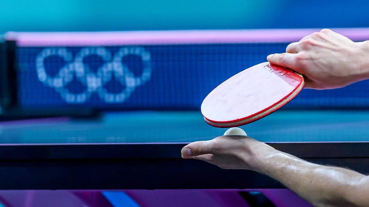 Ein Tischtennisspieler hält seinen Schläger und einen Ball vor der Platte. Im Hintergrund sind die Olympischen Ringe zu erkennen. 