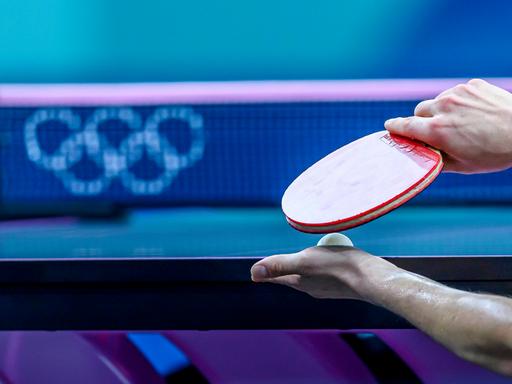 Ein Tischtennisspieler hält seinen Schläger und einen Ball vor der Platte. Im Hintergrund sind die Olympischen Ringe zu erkennen. 