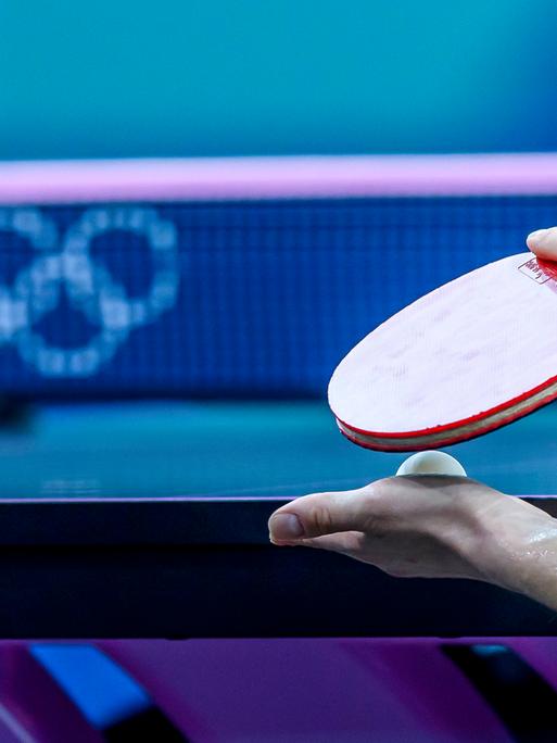 Ein Tischtennisspieler hält seinen Schläger und einen Ball vor der Platte. Im Hintergrund sind die Olympischen Ringe zu erkennen. 