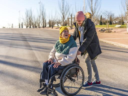 Junger Pfleger hilft einer fröhlichen älteren Frau im Rollstuhl, die einen sonnigen Wintertag in einem Park genießt 
