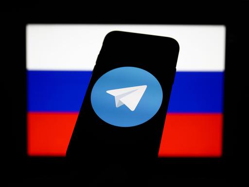Das Logo des Messengerdienstes Telegram auf dem Bildschirm eines Mobiltelefons, eingebettet in die russische Fahne. 