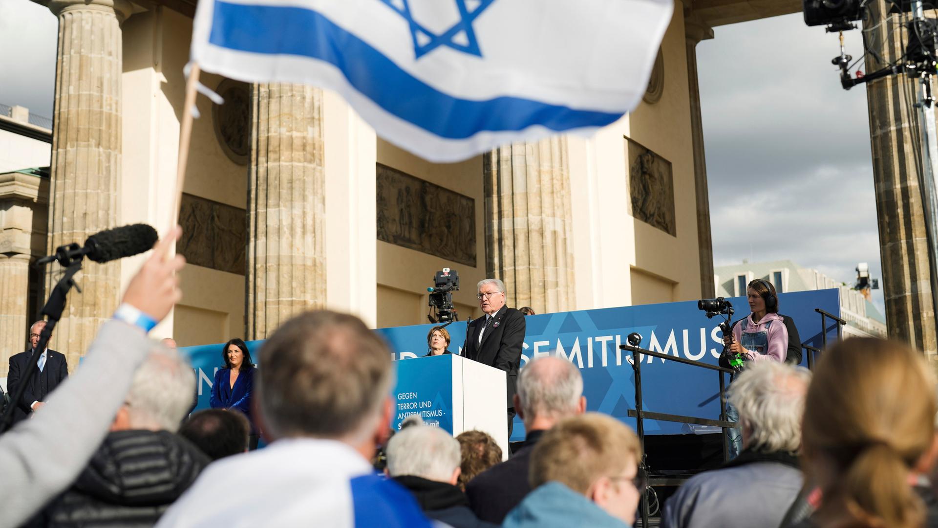 Das Foto zeigt Bundespräsident Steinmeier bei einer Demonstration am Brandenburger Tor in Berlin. Menschen schwenken eine große israelische Flagge.