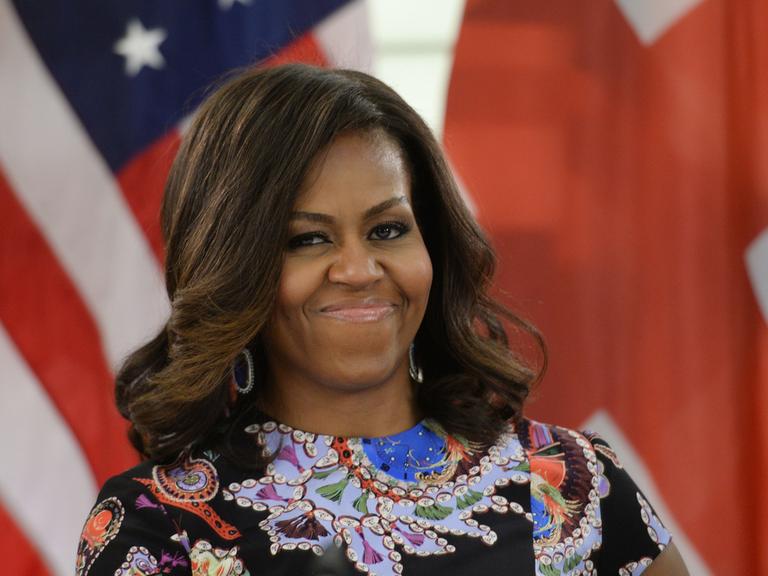 Michelle Obama steht lächeln vor einer amerikanischen Flagge.