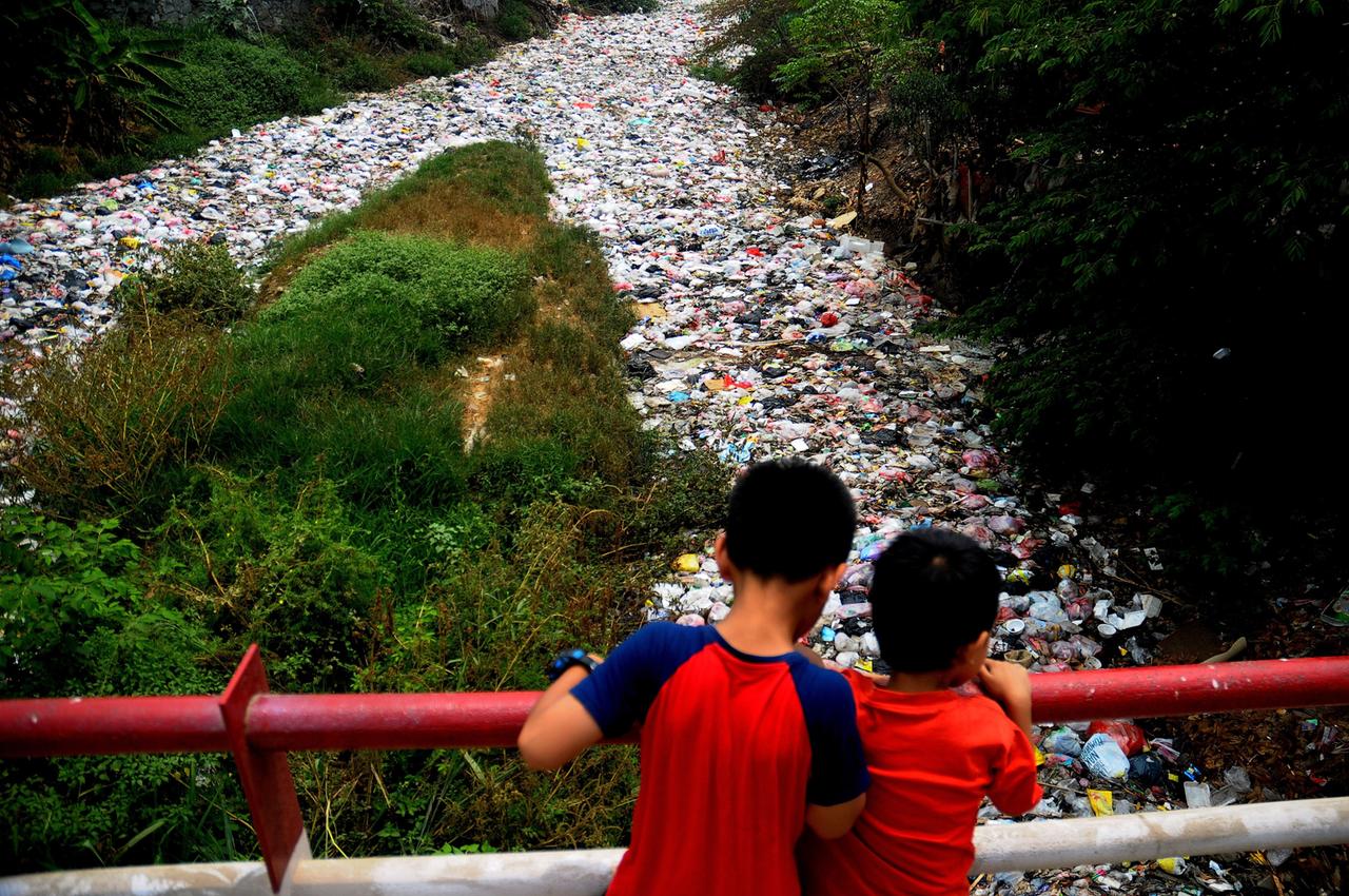 Das Bild zeigt zwei Kinder, die von einer Brücke aus auf einen Fluss voller Plastikmüll schauen Das Bild zeigt zwei Kinder, die von einer Brücke aus auf einen Fluss voller Plastikmüll schauen