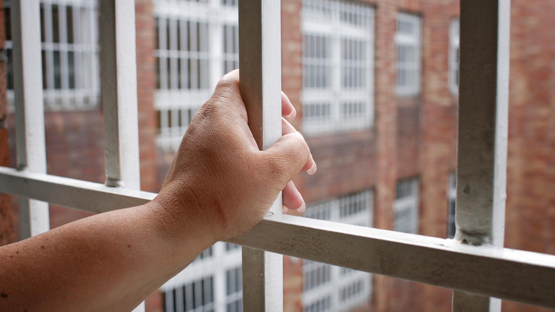 Ersatzfreiheitsstrafen werden seit Jahren kontrovers diskutiert. Zu sehen: Eine Hand umfasst das Gitter an einem Gefängnisfenster mit Blick auf einen Innenhof