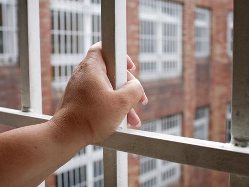 Ersatzfreiheitsstrafen werden seit Jahren kontrovers diskutiert. Zu sehen: Eine Hand umfasst das Gitter an einem Gefängnisfenster mit Blick auf einen Innenhof