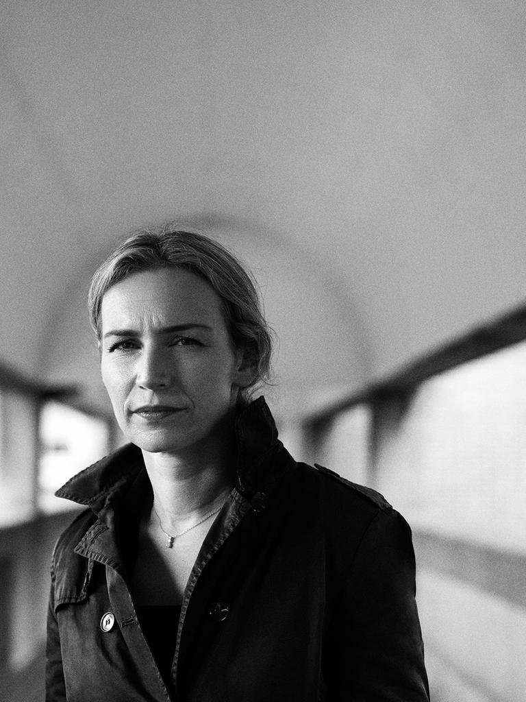 Die Autorin Lea Ypi auf einen Schwarzweiß-Foto. Sie steht in einem langen Gang mit Fenstern auf beiden Seiten.