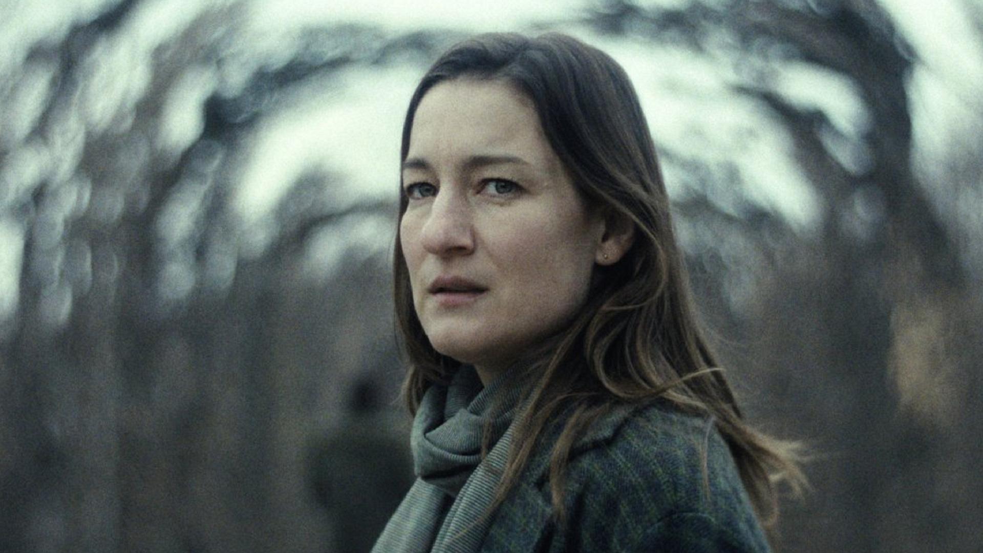 Eine Frau mit langen braunen Haaren schaut verängstigt in die Kamera, im Hintergrund verschwommene Bäume.