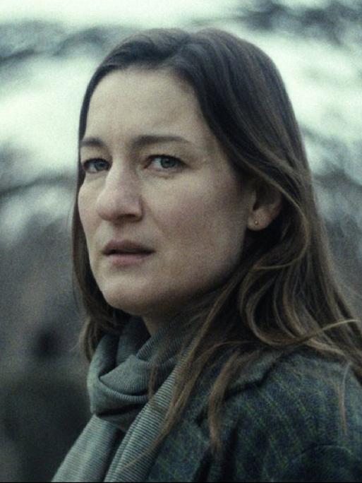 Eine Frau mit langen braunen Haaren schaut verängstigt in die Kamera, im Hintergrund verschwommene Bäume.