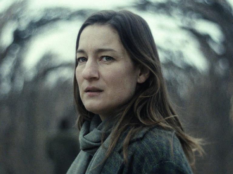 Eine Frau mit langen braunen Haaren schaut verängstigt in die Kamera, im Hintergrund verschwommene Bäume.