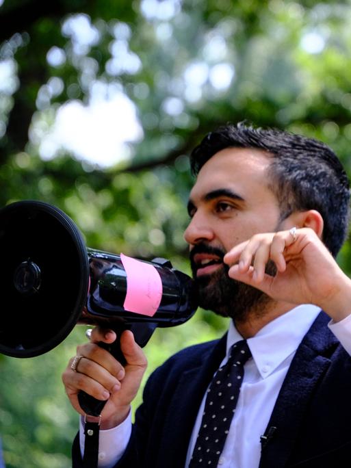 Ein Mann mit kurzen schwarzen Haaren und Bart im Anzug spricht in ein Megaphon. Ein Mann mit kurzen schwarzen Haaren und Bart im Anzug spricht in ein Megaphon.