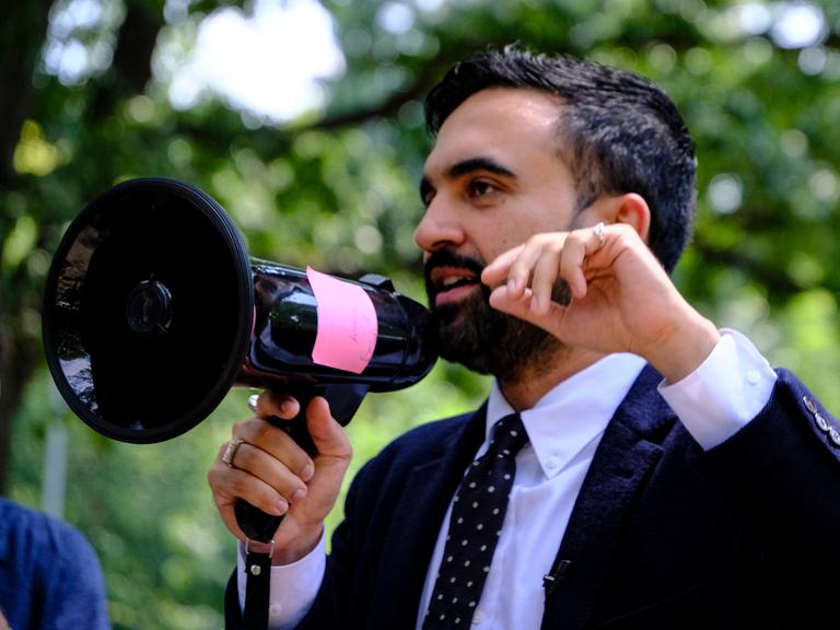 Ein Mann mit kurzen schwarzen Haaren und Bart im Anzug spricht in ein Megaphon. Ein Mann mit kurzen schwarzen Haaren und Bart im Anzug spricht in ein Megaphon.