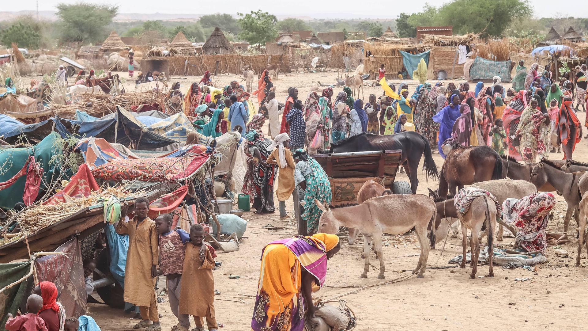 Aus dem Sudan geflüchtete Menschen im Tschad. Zu sehen sind vor allem Frauen und Kinder, die vor Lehmhütten und neben provisorischen Unterständen stehen.