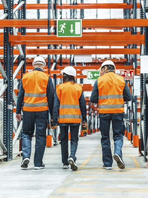 Lagerarbeiter, die Warnwesten und Schutzhelme tragen, gehen durch ein Logistikzentrum 