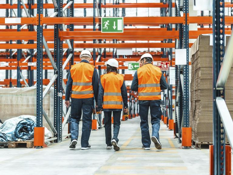 Lagerarbeiter, die Warnwesten und Schutzhelme tragen, gehen durch ein Logistikzentrum 