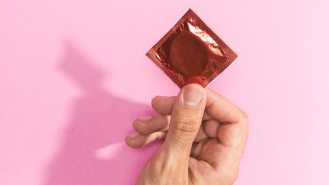 Eine Hand hält eine geschlossene Kondompackung vor einem rosa Hintergrund.