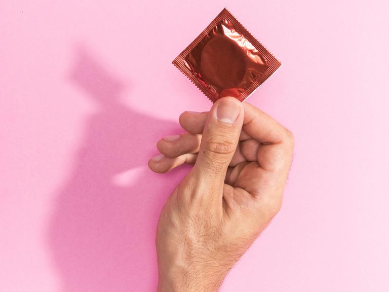 Eine Hand hält eine geschlossene Kondompackung vor einem rosa Hintergrund.