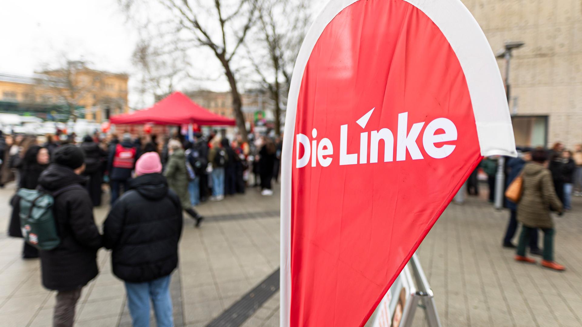 Eine Fahne mit dem Schriftzug "Die Linke" steht an einem Infostand der Partei in einer Innenstadt, im Hintergrund sind Menschen zu sehen.