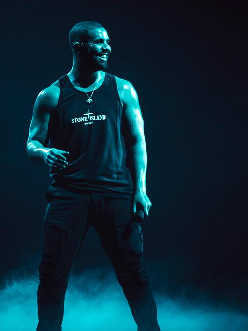 Der kanadische Rapper Drake steht  in schwarzem Hemd auf einer Bühne. Der Hintergrund in dunkel.