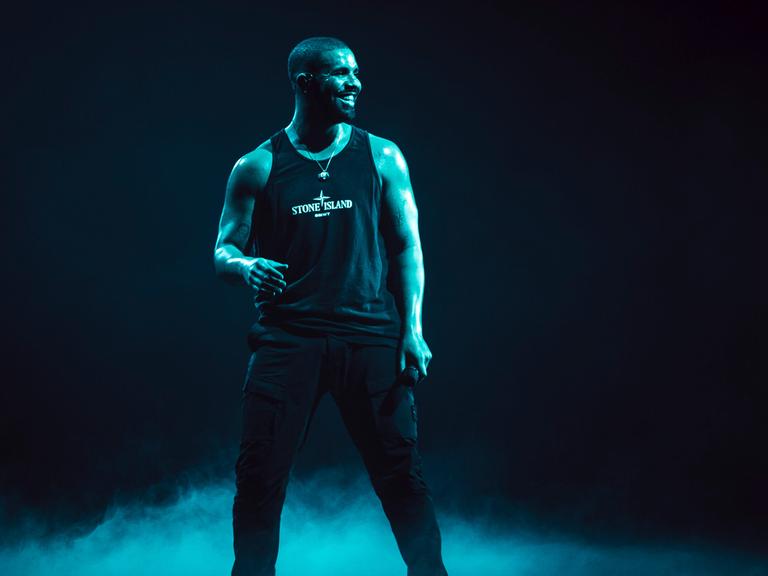Der kanadische Rapper Drake steht  in schwarzem Hemd auf einer Bühne. Der Hintergrund in dunkel.