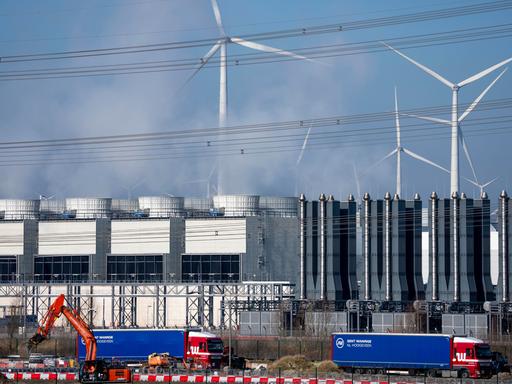 Ein Rechenzentrum, man sieht Kühltürme der Serverkühlung, Windräder und im Vordergrund Bagger und LKW auf einer Baustelle.