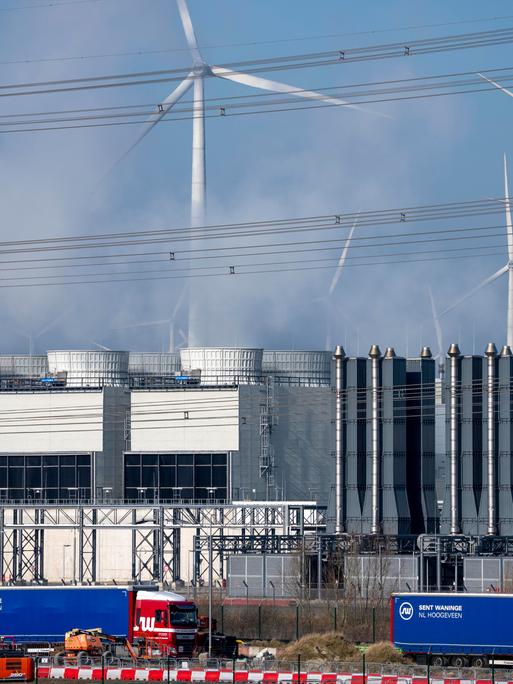 Ein Rechenzentrum, man sieht Kühltürme der Serverkühlung, Windräder und im Vordergrund Bagger und LKW auf einer Baustelle.