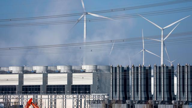 Ein Rechenzentrum, man sieht Kühltürme der Serverkühlung, Windräder und im Vordergrund Bagger und LKW auf einer Baustelle.