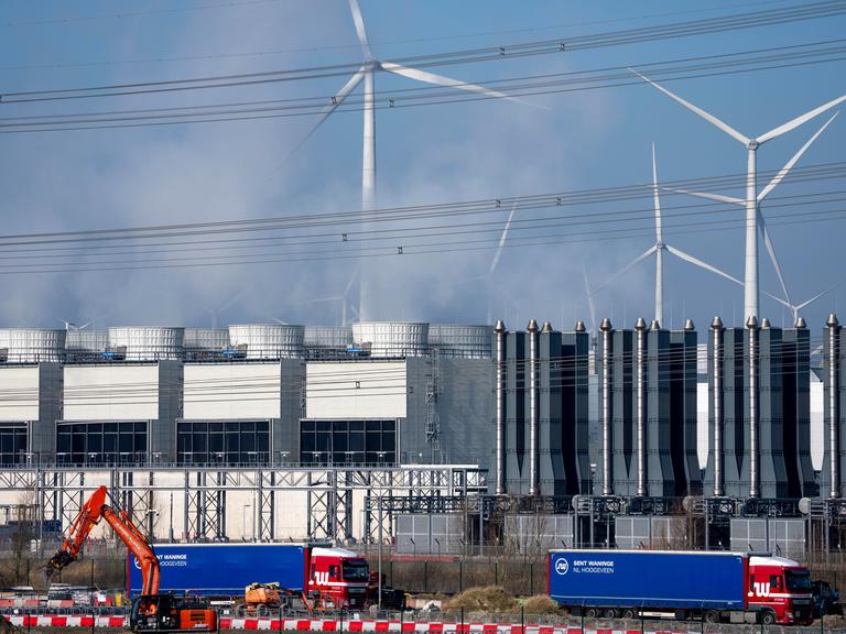 Ein Rechenzentrum, man sieht Kühltürme der Serverkühlung, Windräder und im Vordergrund Bagger und LKW auf einer Baustelle.