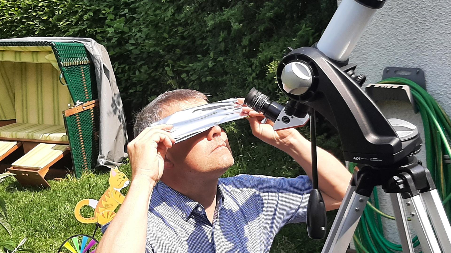 Ein Mann hält eine gefaltete Folie mit beiden Händen über seine Augen und schaut in Richtung Himmel. Neben ihm steht ein Teleskop.